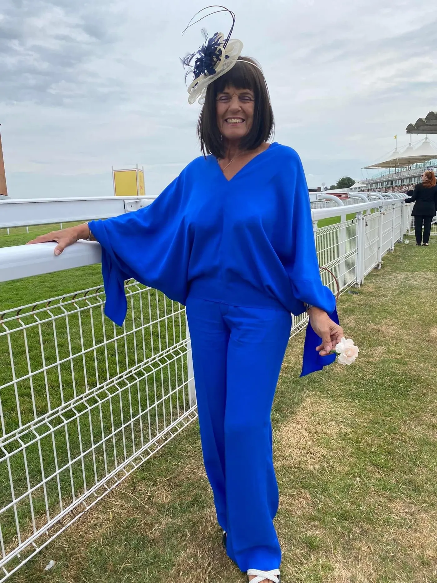 Linda, dressed in blue, smiling beside the rail on a sunny day at the races